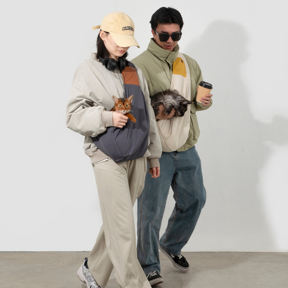 Two people walking side by side, each carrying a cat chest-high in a ComfyPaws shoulder sling; the man on the right also holds a takeaway coffee