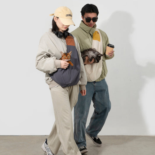 Two people walking side by side, each carrying a cat chest-high in a ComfyPaws shoulder sling; the man on the right also holds a takeaway coffee