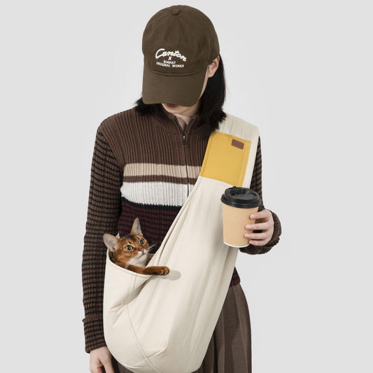 Person wearing a cream cotton sling across the body, the strap over one shoulder and the pouch at the opposite hip, with an Abyssinian cat's head and one front paw out of the pouch and a coffee cup in one hand