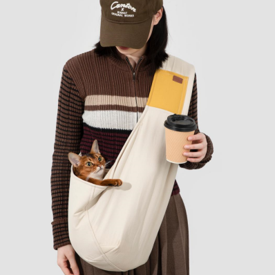 Person holding a coffee cup and a cat in a sling, wearing a brown cap and striped shirt.
