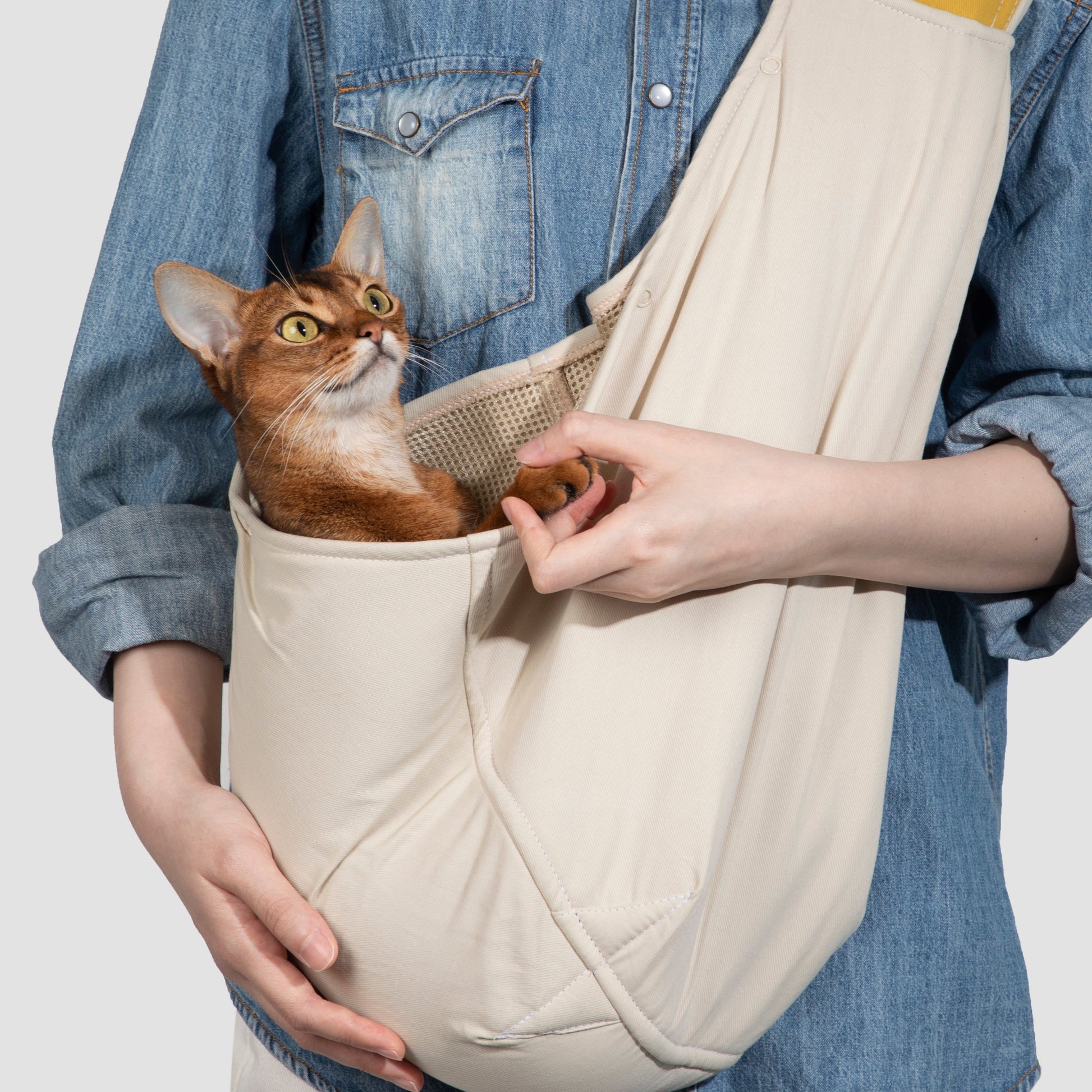 Person holding a beige pet sling with a cat inside on a plain background